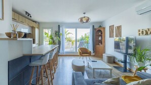Eine moderne Küche mit Barhocker, einem Esstisch und einem Fernseher. Im Wohnbereich befindet sich eine Couch und ein Sessel, sowie eine Pflanze rechts.