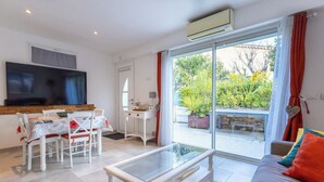 Una sala de estar moderna con un sofá, una mesa de centro de vidrio, una mesa de comedor con sillas, un televisor y un gran ventanal corredizo que da a un balcón con plantas.
