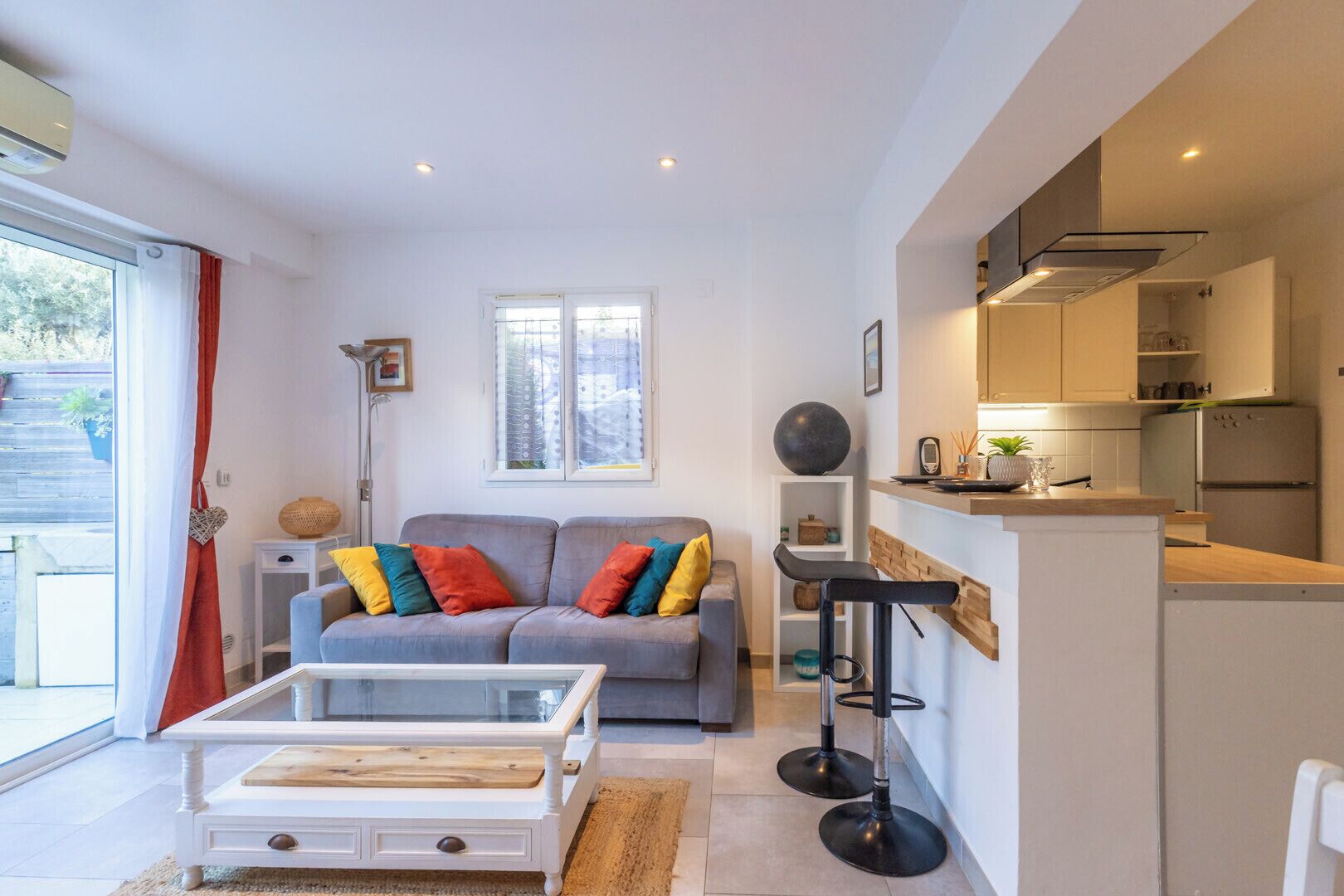 Ein modernes Wohnzimmer mit grauem Sofa, bunten Kissen, einem weißen Couchtisch und einer Küchenzeile mit Einbauherd und Kühlschrank.