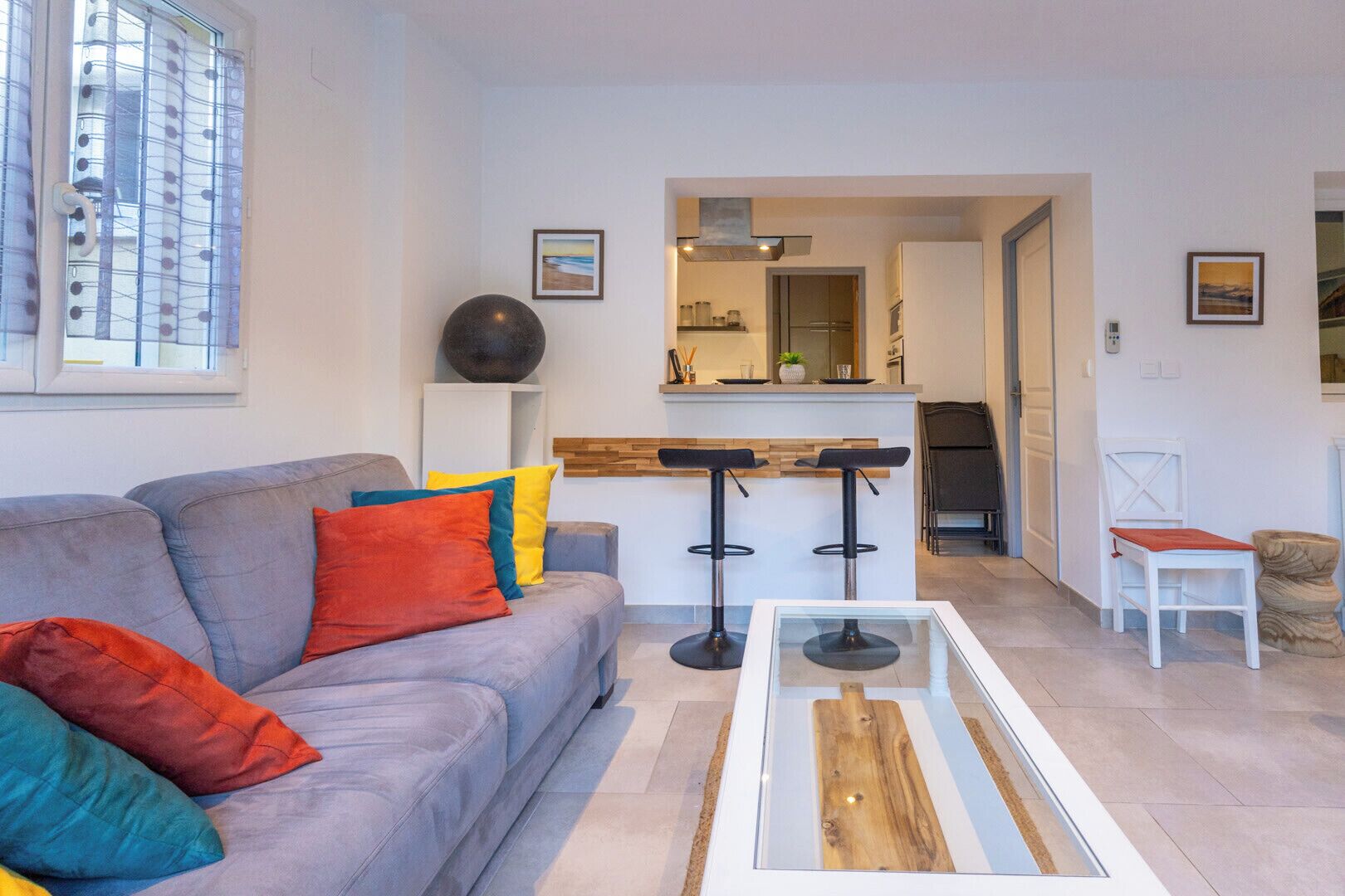A modern living room with a grey sofa, colorful cushions, a glass coffee table, and a kitchen area in the background.