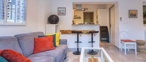 A modern living room with a grey sofa, colorful cushions, a glass coffee table, and a kitchen area in the background.
