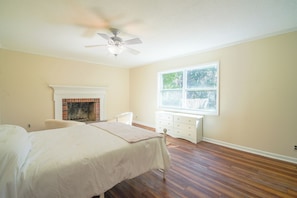 A bedroom with a bed, a white dresser, a fireplace, and a window with a view of trees. - The Marsh House (Brunswick)