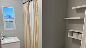 Ein Badezimmer mit Toilette, Waschbecken und Duschvorhang. Über dem Waschbecken befindet sich ein Fenster und eine Ablage mit Toilettenpapier.