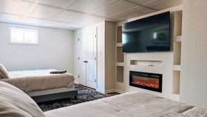 Une chambre d’hôtel moderne dotée d’un grand téléviseur à écran plat fixé au mur, d’une cheminée, de deux lits avec une literie de couleur beige et d’une fenêtre laissant entrer la lumière naturelle.
