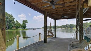 A wooden deck with a hanging hammock and a ceiling fan, overlooking a calm lake with trees and a distant shoreline. - Well-appointed 5-bedroom, 5 bathroom house on gorgeous Lake Wateree with AC (Ridgeway)