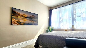 Un dormitorio con cama, una ventana con cortinas, un cuadro en la pared y una planta.