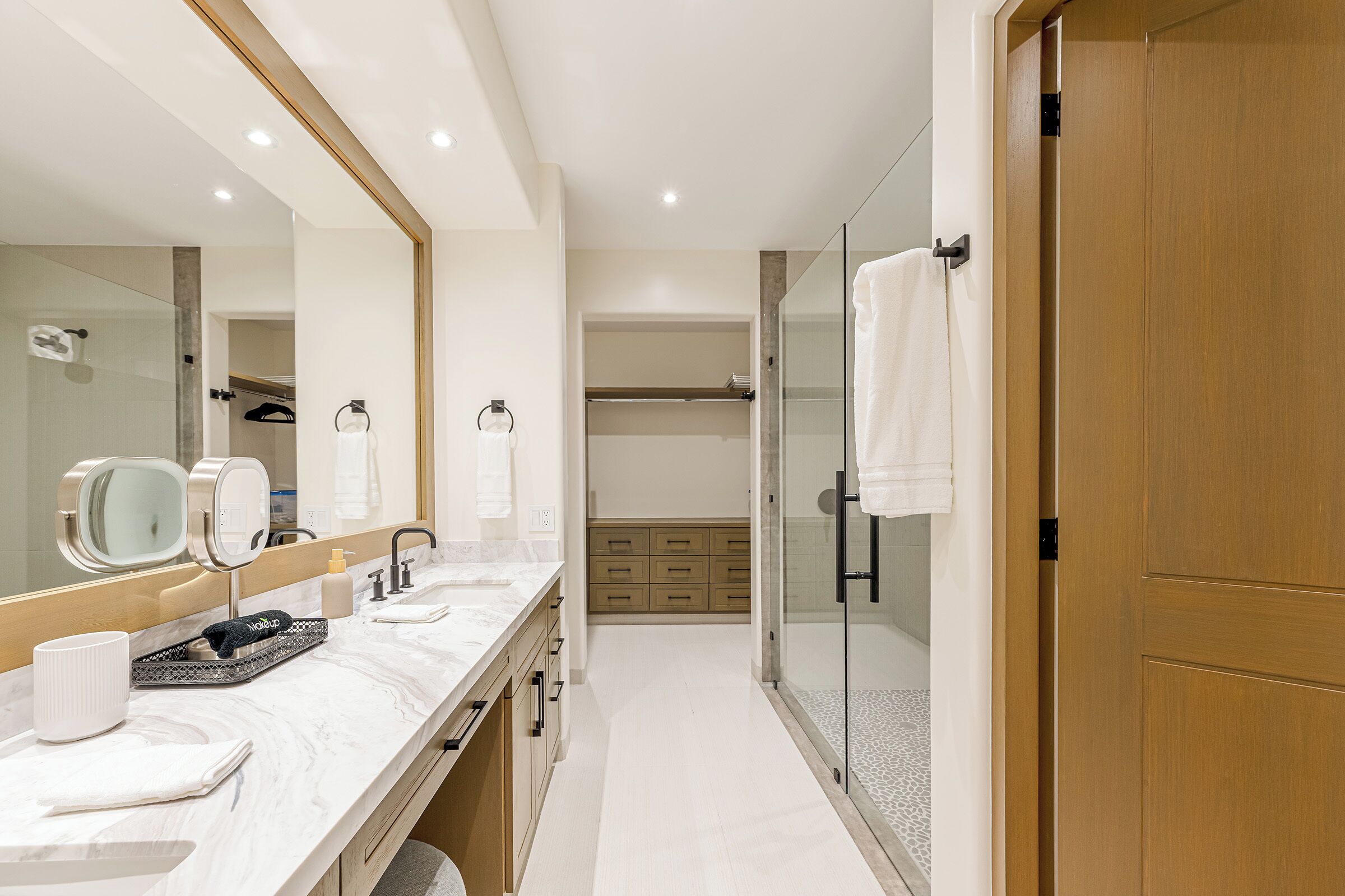 A modern bathroom with a large mirror, two sinks, a shower, and a walk-in closet.