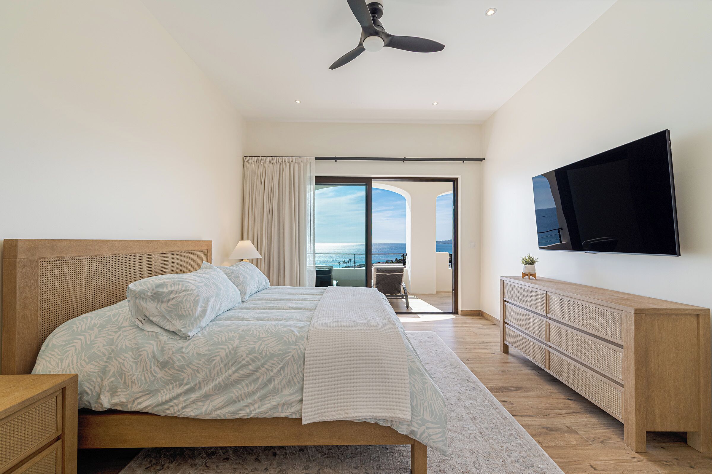 A modern bedroom with a large bed, a ceiling fan, a TV, and a view of the sea through a sliding door.