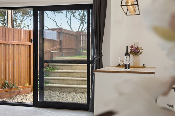 A modern interior with a glass door leading to a wooden deck, a small table with a bottle and glass, and a wall-mounted light fixture.