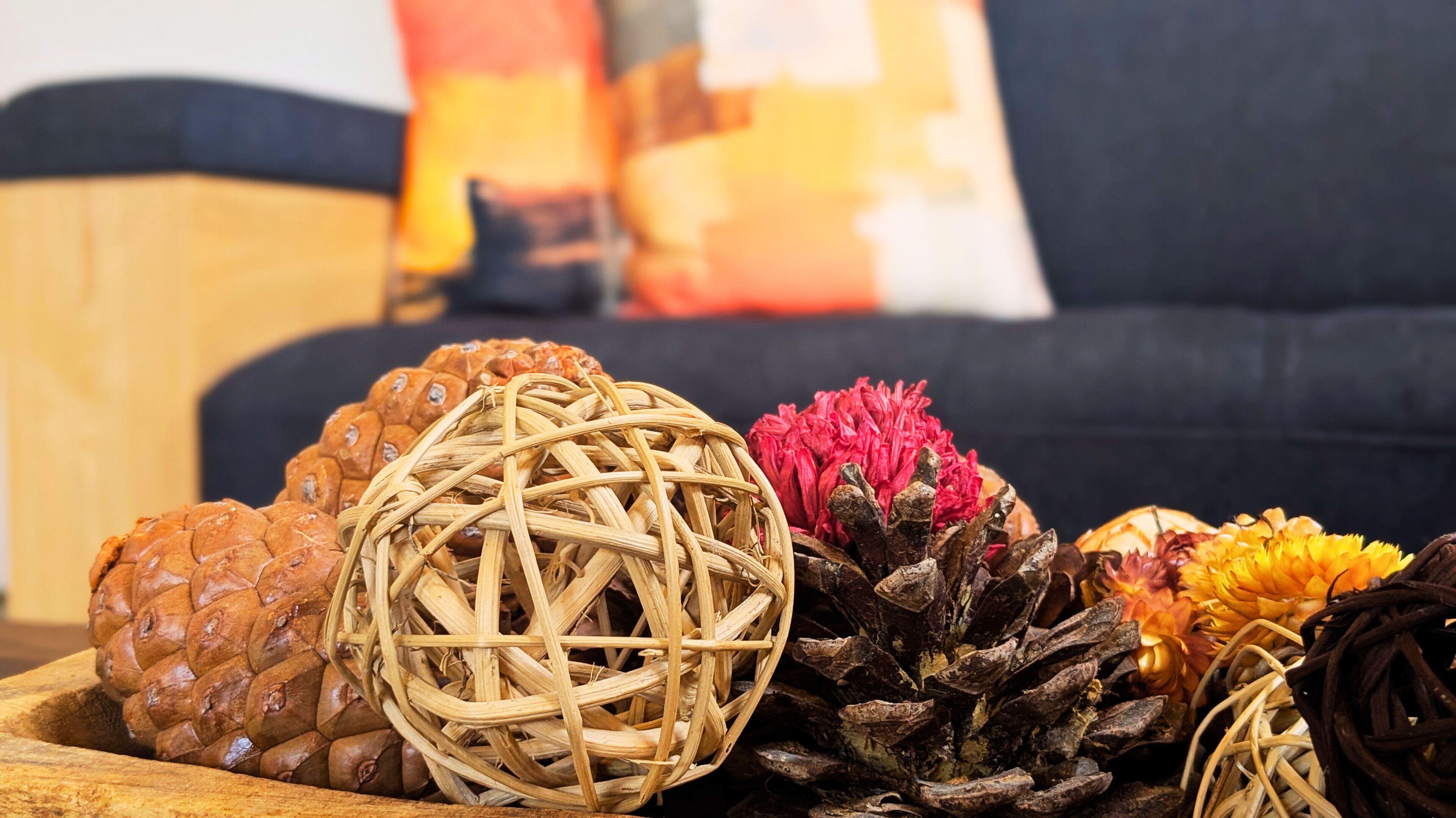 Eine dekorative Anordnung aus Naturmaterialien, darunter eine Kiefernzapfen, eine gewebte Kugel und getrocknete Blumen auf einer Holzoberfläche.
