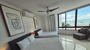 Moderni hotellihuone, jossa on kaksi sänkyä, kattotuuletin ja suuri ikkuna, josta avautuu näkymä merelle.