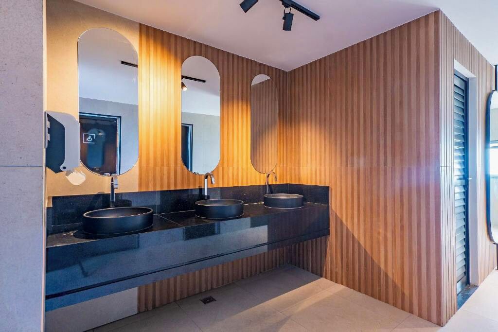 Un baño moderno con paneles de madera, tres lavamanos negros y espejos grandes.