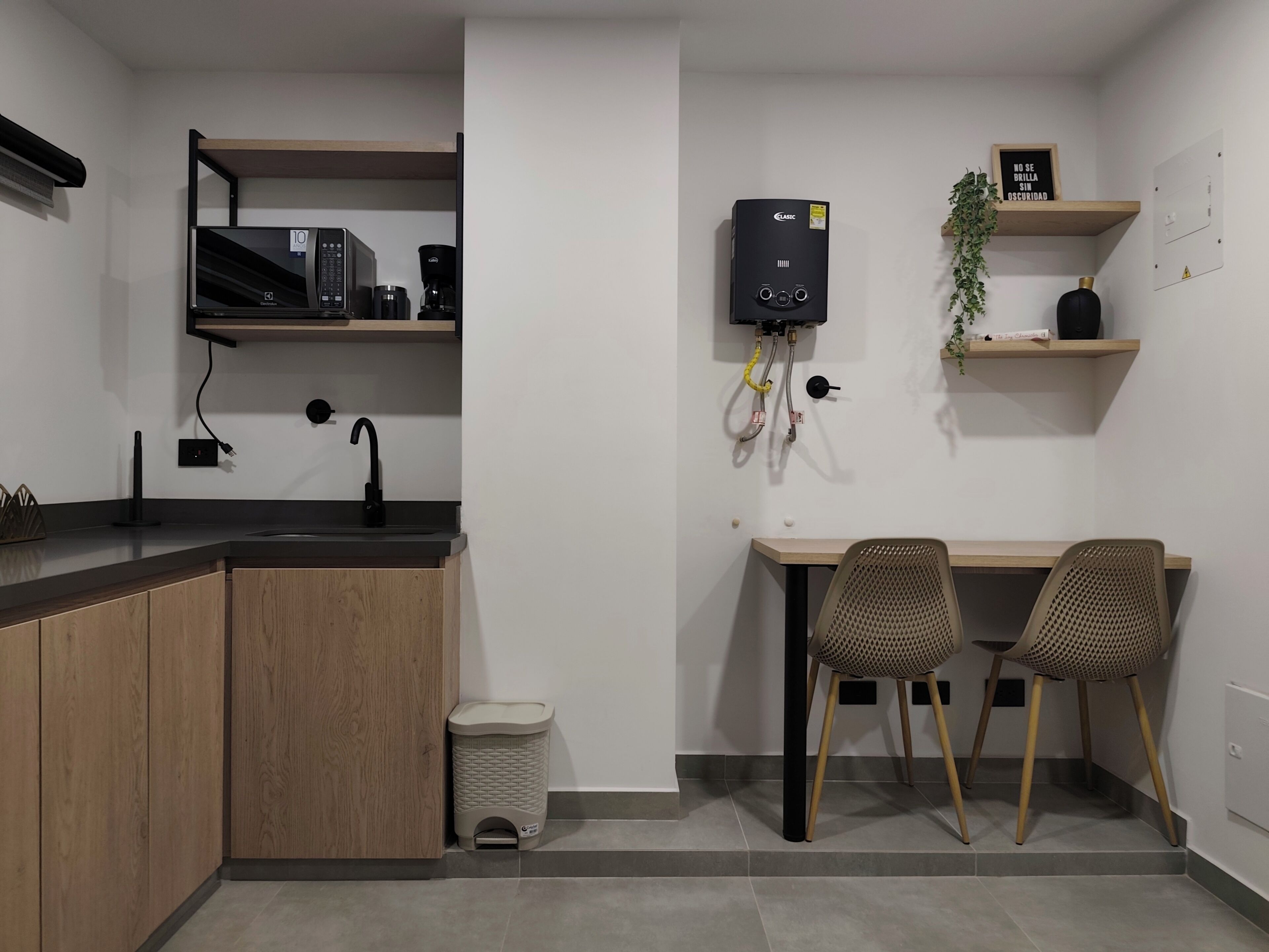 A compact kitchen with a sink, countertop, and a coffee machine. There is a trash bin, a small table with two chairs, and a heater mounted on the wall.