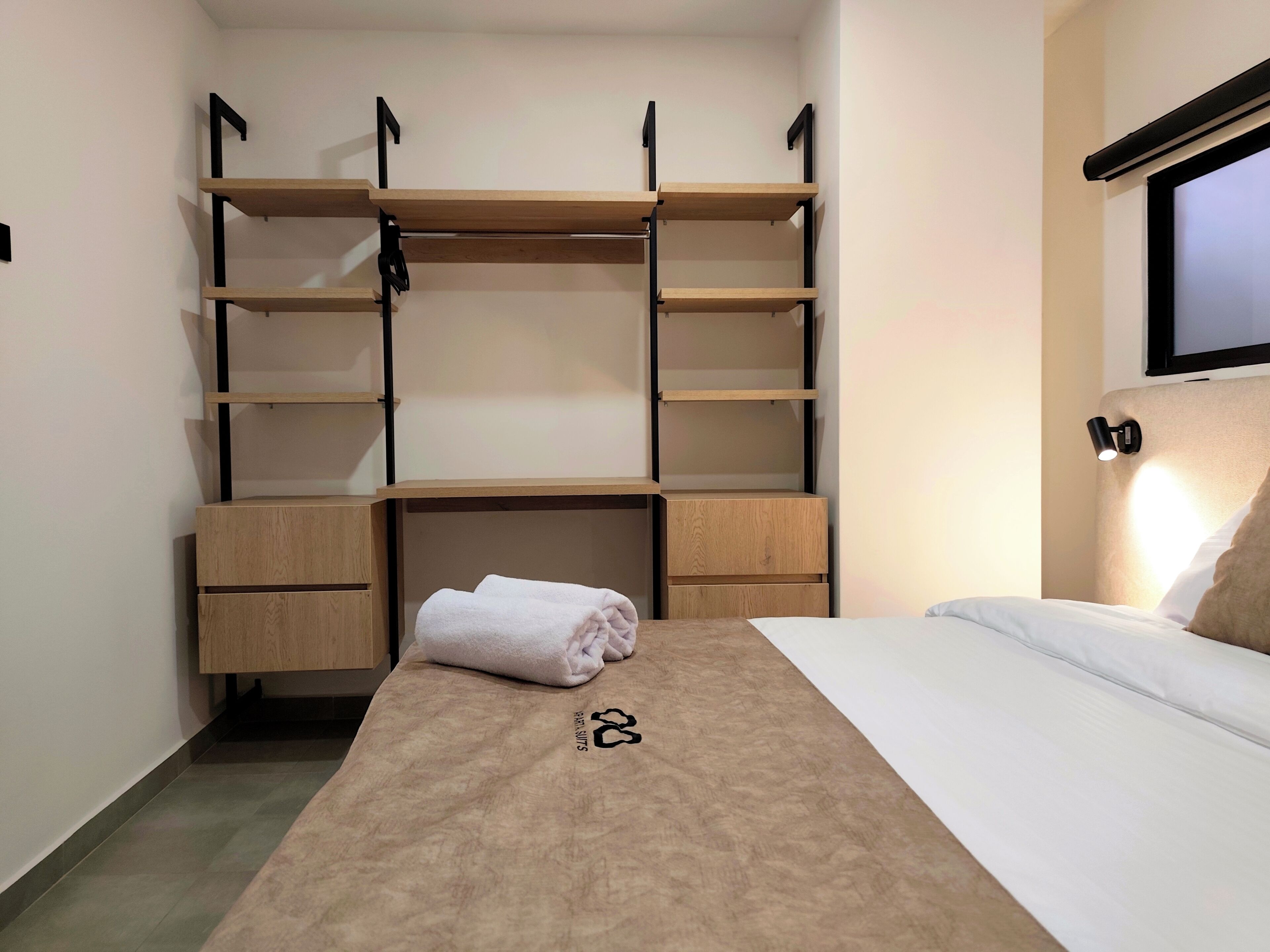 Un dormitorio con cama, armario empotrado con estantes múltiples, mesita de noche y televisor montado en la pared.