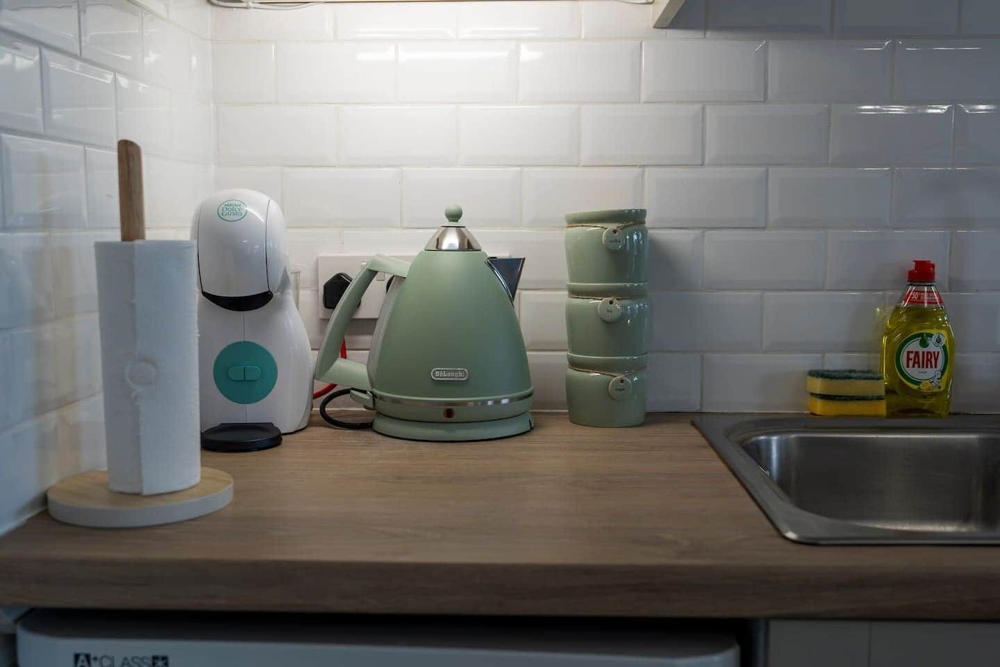 Et køkken med elkedel, kaffemaskine, en stak kopper og en flaske Fairy opvaskemiddel.