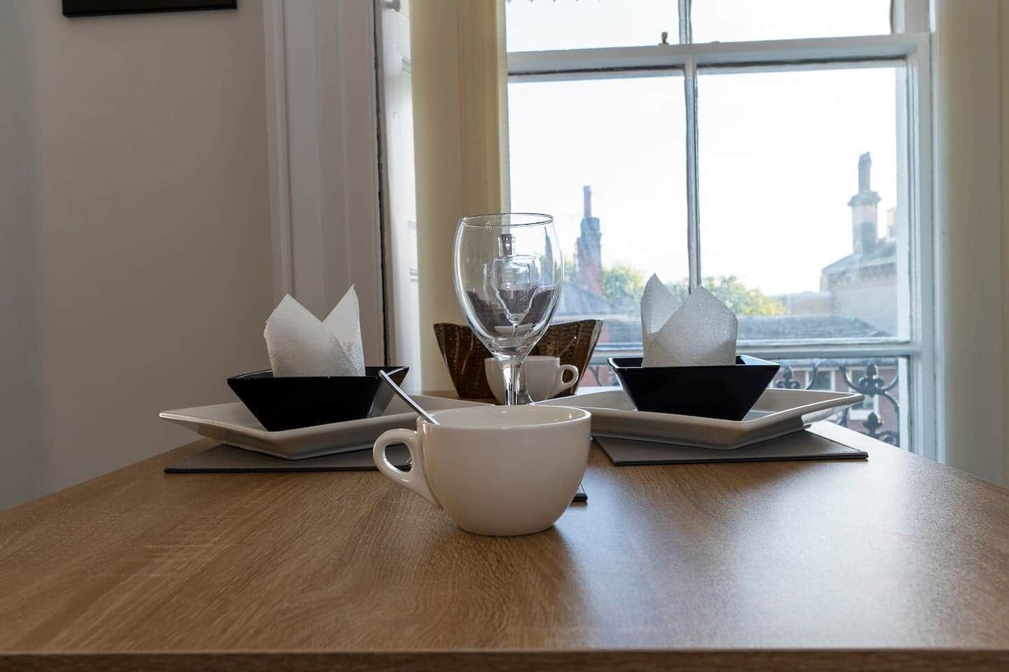 A table with a white cup, a glass, and two black napkin holders with napkins, set against a window with a view of buildings.