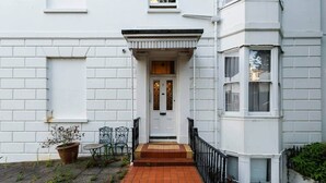 Un bâtiment blanc avec une entrée surmontée de tuiles rouges, une rampe en métal noir et un petit jardin agrémenté de plantes en pot.