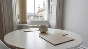 Une table ronde blanche, sur laquelle se trouvent un vase blanc et deux cahiers, dans une pièce aux murs blancs et dotée d’une fenêtre donnant sur un immeuble.