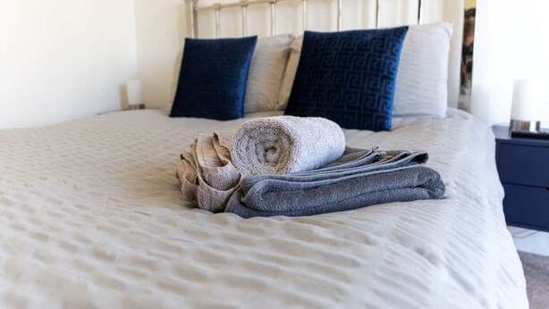 A neatly made bed with a beige bedspread, two rolled-up towels, and two pillows on a headboard.