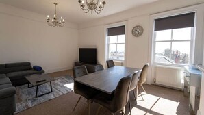 A living room with a sofa, coffee table, TV, dining table with chairs, a chandelier, and large windows with blinds.