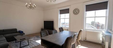 A living room with a sofa, coffee table, TV, dining table with chairs, a chandelier, and large windows with blinds.