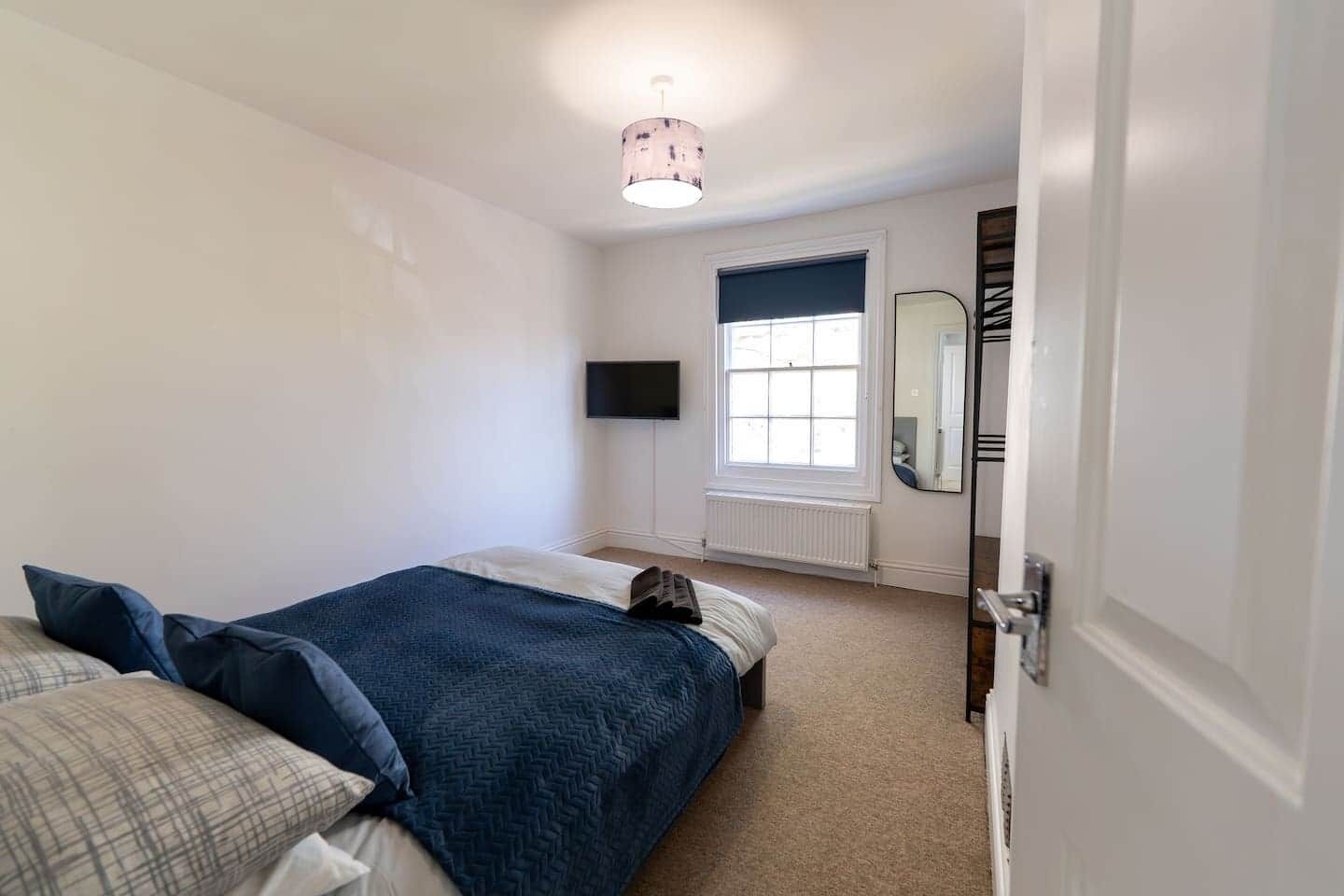 A bedroom with a bed, a window, a mirror, and a wall-mounted TV.
