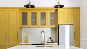 A kitchen with yellow cabinets, a white countertop, a stainless steel refrigerator, and three white stools.