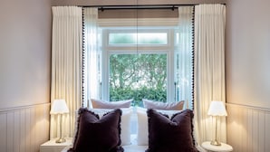 A bedroom with a large bed, two dark pillows, white curtains, and a window with a view of greenery.