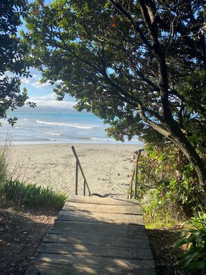 Un sendero de madera que conduce a una playa arenosa, con vista despejada al océano y montañas distantes.