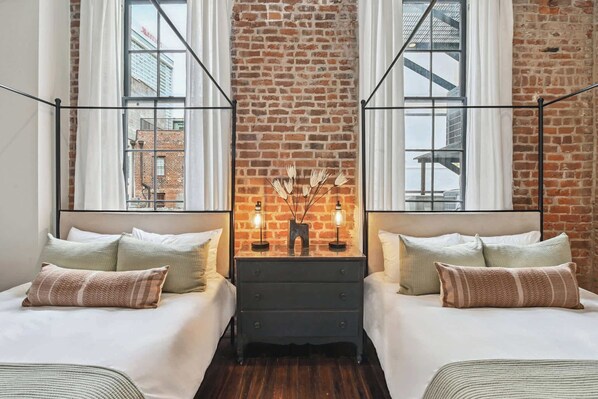 A room with two beds, a brick wall, and a view of buildings through large windows.