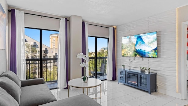 Un salon moderne avec un canapé gris, une petite table ronde, un téléviseur fixé au mur et un balcon agrémenté de rideaux violets.