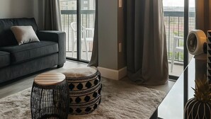 A modern living room with a grey sofa, a round wooden side table, a patterned ottoman, and a large window with curtains. - 3704 Fully remodeled & modern Enclave at Unviersal (Orlando)