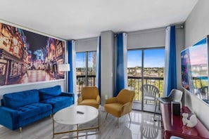 A modern hotel room with a large cityscape painting, a blue sofa, a round table, and a balcony with a view. - 3805 The Enclave Orlando: A Magical Retreat (orlando)