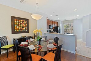 A dining area with a glass table, chairs, and a vase of flowers. There is a kitchen in the background with wooden cabinets and a microwave. - 5026-13 Spacious 3BR Townhome at Vista Cay Resort (Orlando)