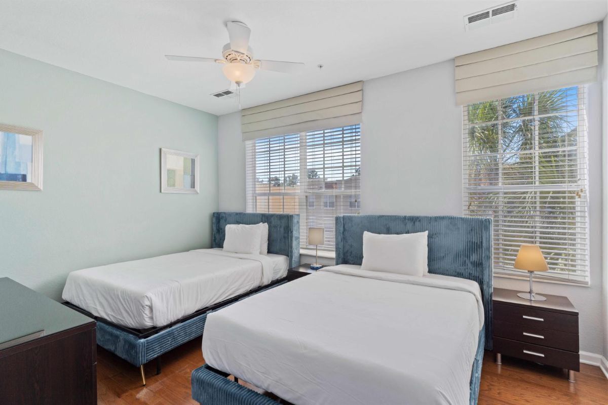 A hotel room with two beds, a ceiling fan, a window with blinds, and a nightstand with a lamp.