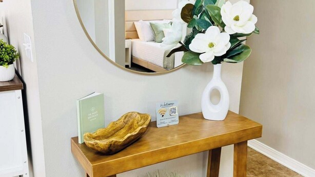 Ein Holzkonsolentisch mit Spiegel, einer Willkommenskarte, einem Buch und einer Vase mit Blumen.