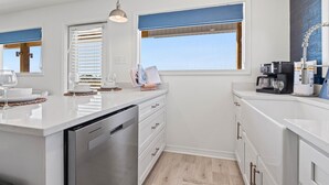 A modern kitchen with white cabinetry, a dishwasher, a coffee maker, and a window with blinds. - High Point Hideaway on the Sand Dunes - Unit 2 (Dauphin Island)
