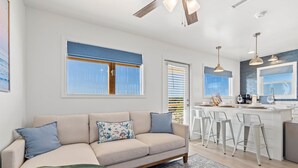 A modern living room with a sofa, a coffee table, and a patterned rug. There is a kitchen area with a bar and stools. - High Point Hideaway on the Sand Dunes - Unit 2 (Dauphin Island)