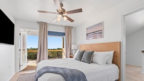 A bedroom with a bed, a ceiling fan, a nightstand, a lamp, a painting, and a view of the outdoors. - High Point Hideaway on the Sand Dunes - Unit 2 (Dauphin Island)