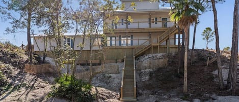 Un edificio de dos pisos con una escalera de madera que lleva a una terraza, rodeado de árboles y terreno arenoso.