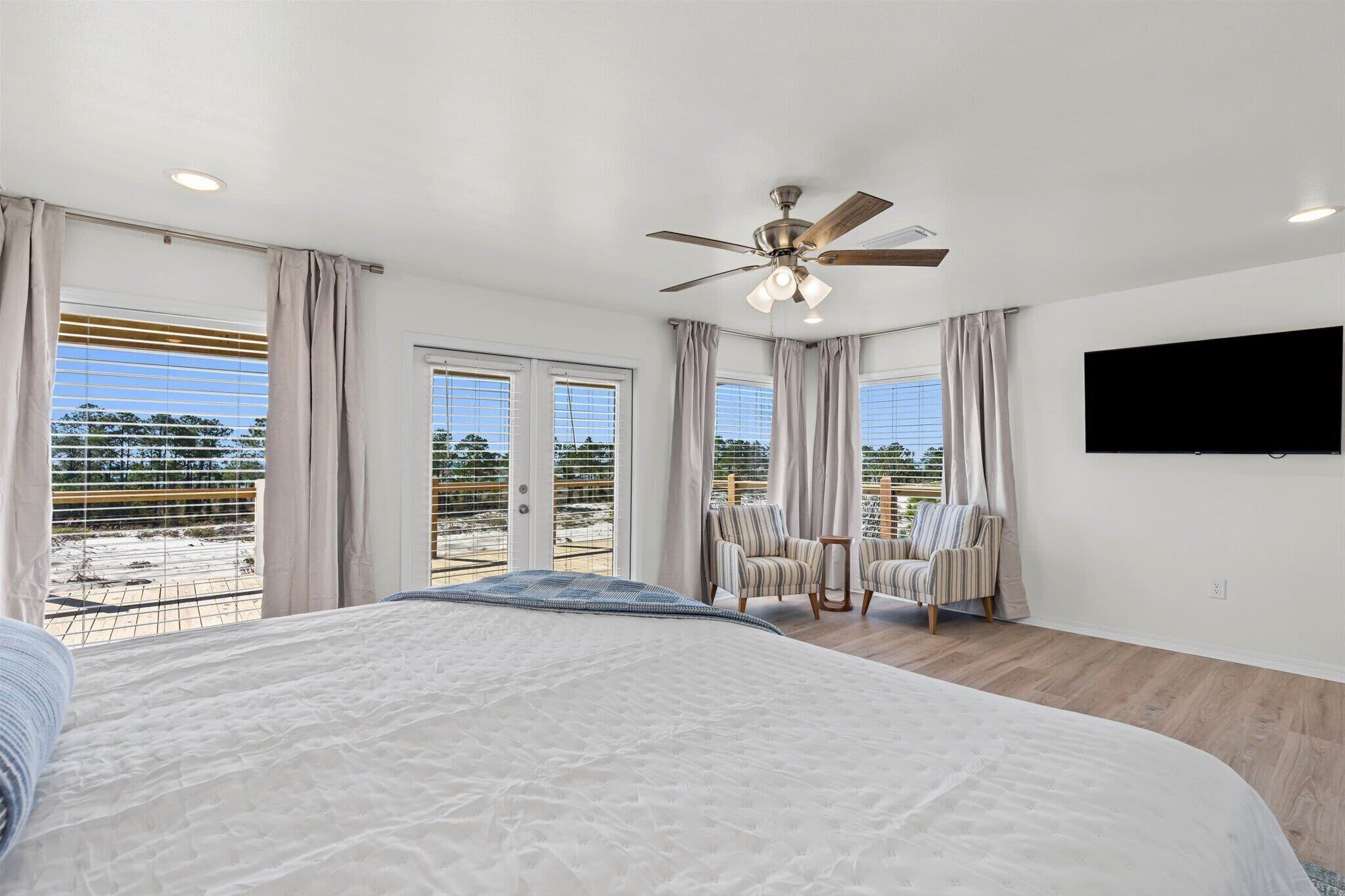 A bedroom with a large bed, a ceiling fan, a TV mounted on the wall, and a balcony with a view of the beach.
