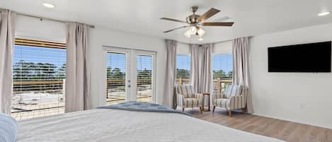 A bedroom with a large bed, a ceiling fan, a TV mounted on the wall, and a balcony with a view of the beach.