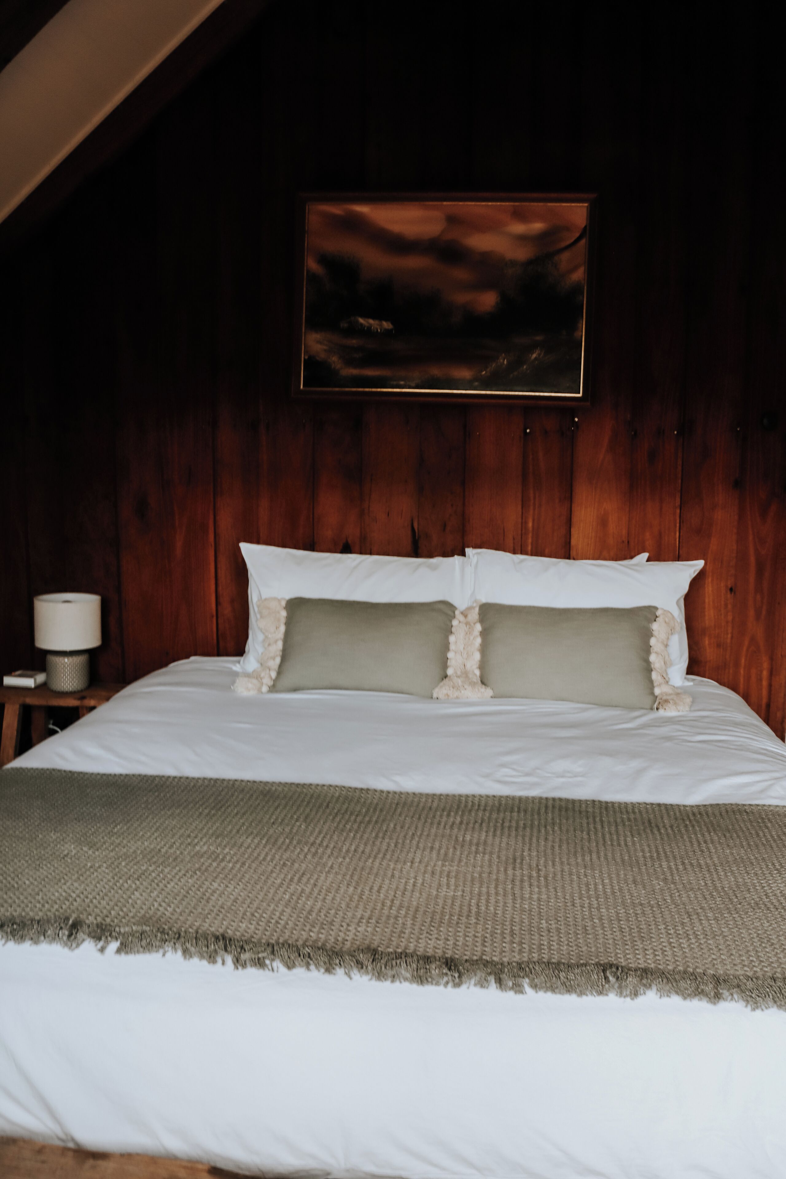 A bedroom with a wooden headboard, a bed with white linens, two pillows, a bedside lamp, and a framed picture on the wall.