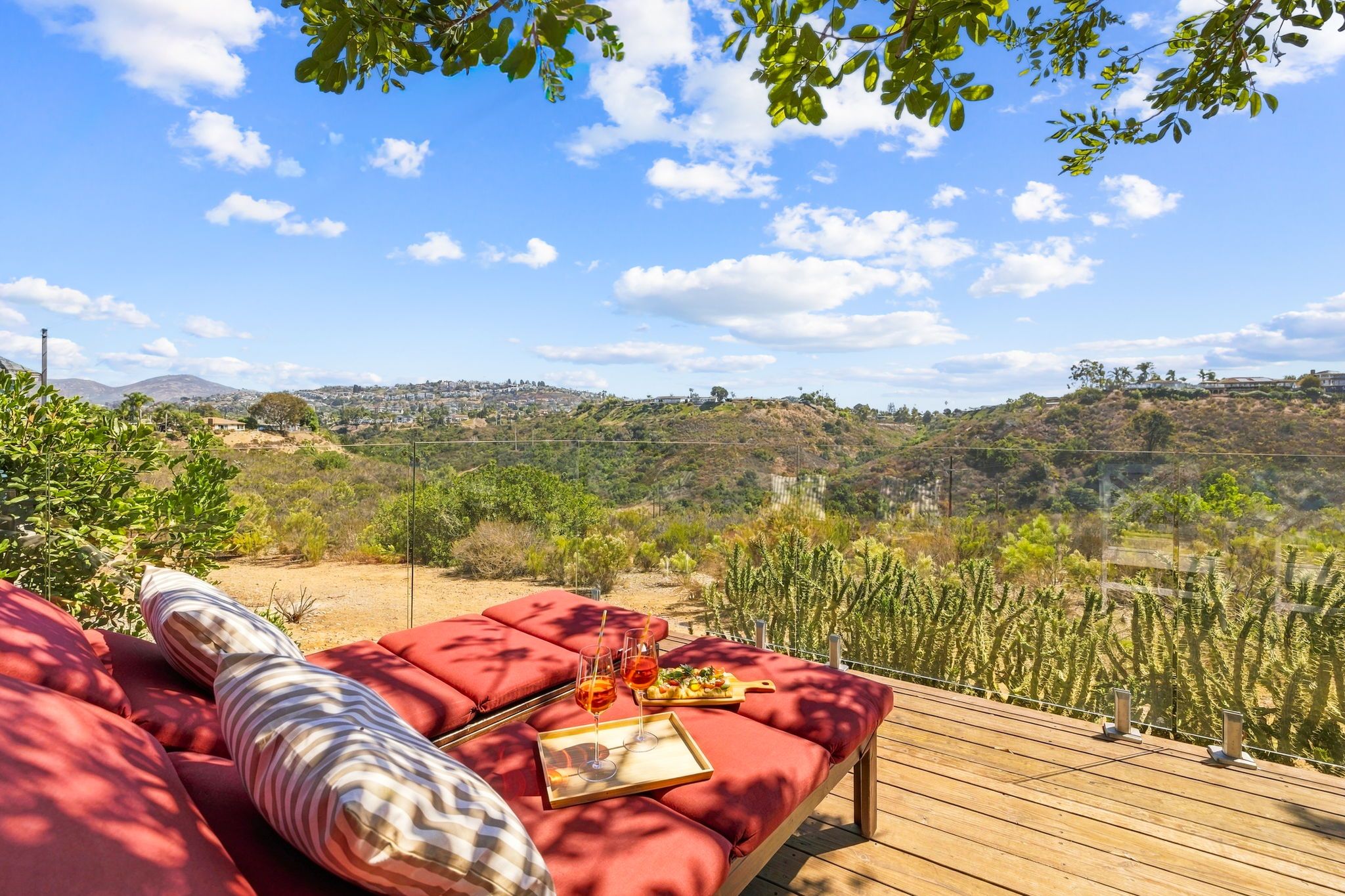 A deck with red cushioned seating, a tray with glasses and a plate of food, and a view of a hilly landscape with vegetation and buildings in the distance.