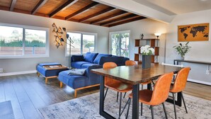 Een ruime woonkamer met een houten plafond, een blauwe bank, oranje fauteuils en een eettafel.