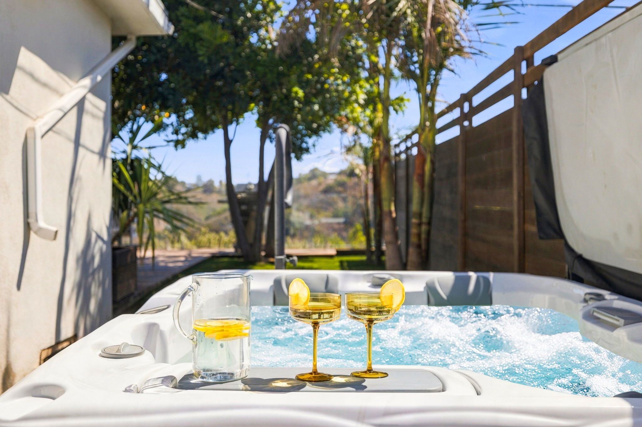 A hot tub with a glass pitcher and two glasses, surrounded by a wooden fence and trees.