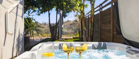 A hot tub with a glass pitcher and two glasses, surrounded by a wooden fence and trees.