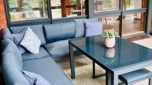 Un área de estar moderna al aire libre con un sofá seccional y una mesa de vidrio, con amplias ventanas que ofrecen vistas al exterior.