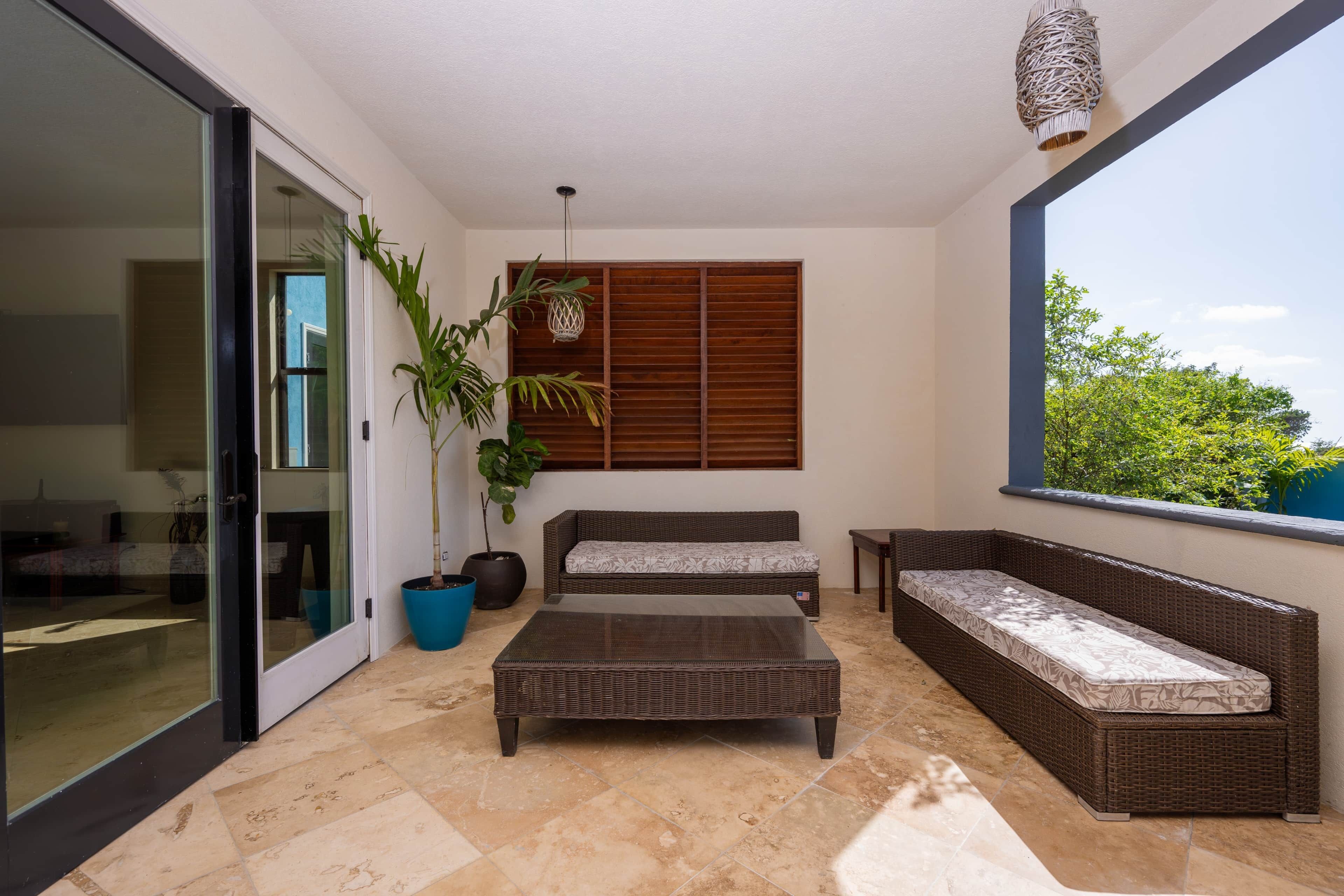 A modern living room with a glass door leading to a balcony, a large potted plant, wicker furniture, and a view of greenery.