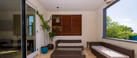 A modern living room with a glass door leading to a balcony, a large potted plant, wicker furniture, and a view of greenery.